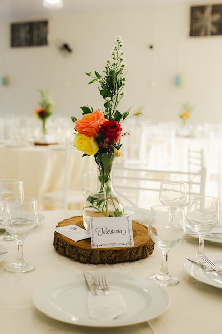A floral arrangement featuring a mix of colorful flowers, including orange roses, a red rose, a yellow bloom, and white and green filler flowers, displayed in a clear glass vase. The bouquet is placed