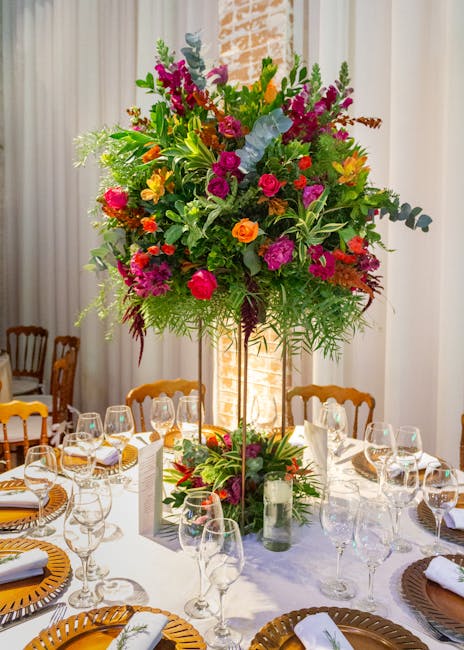 A tall, vibrant floral arrangement featuring a variety of flowers including pink roses, purple delphiniums, orange ranunculus, and blue eucalyptus, all set in a clear glass vase atop a tall gold stand