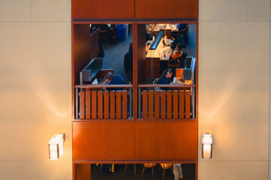 A view through a large window showcasing a modern office environment with several employees working at desks equipped with computer monitors. The workspace features warm lighting, wooden accents, and 