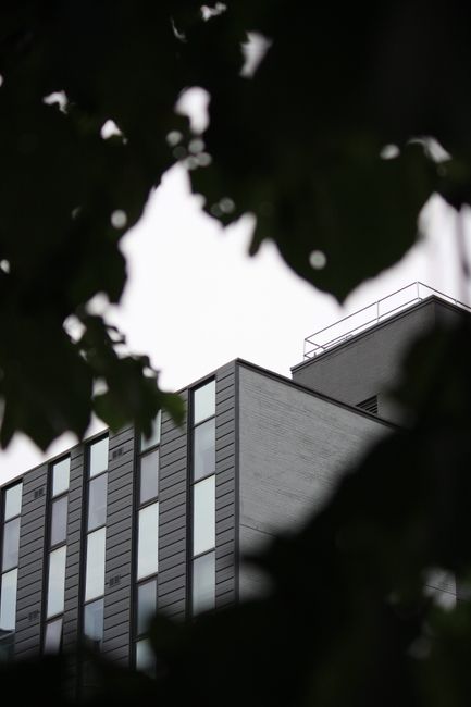 The image shows a modern multi-story building with a sleek exterior composed of dark paneling and large, rectangular windows. The building's clean lines and minimalist design are visible, with a porti