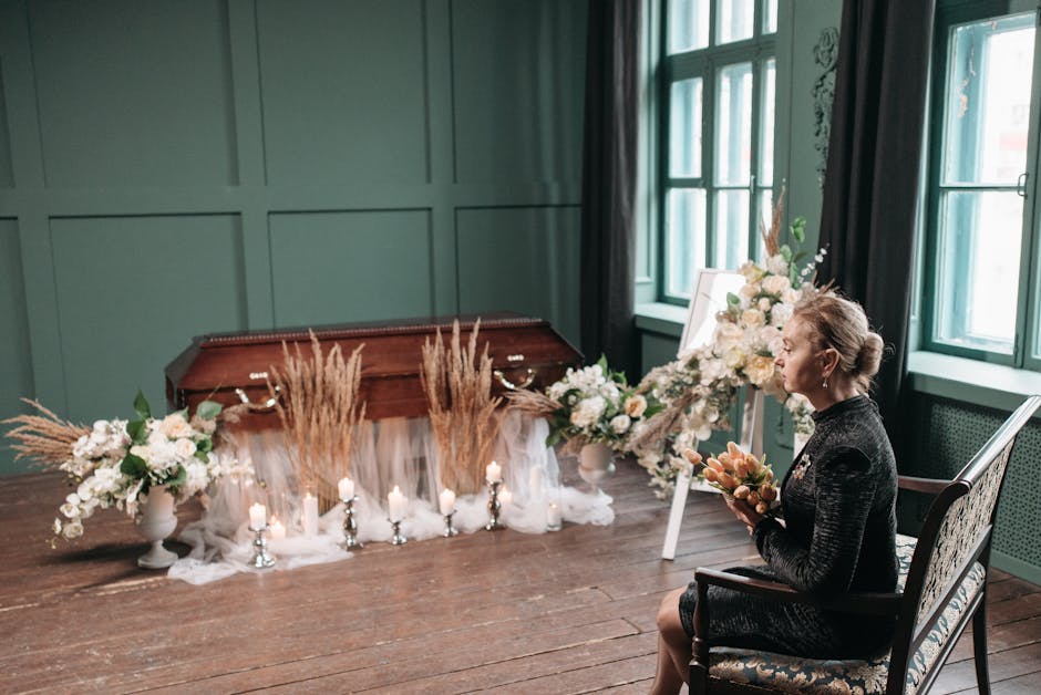 A woman seated indoors holding a floral arrangement in her hands, with an elegant, dark-colored dress and her hair styled in an updo. Behind her, there is a display of large floral arrangements featur