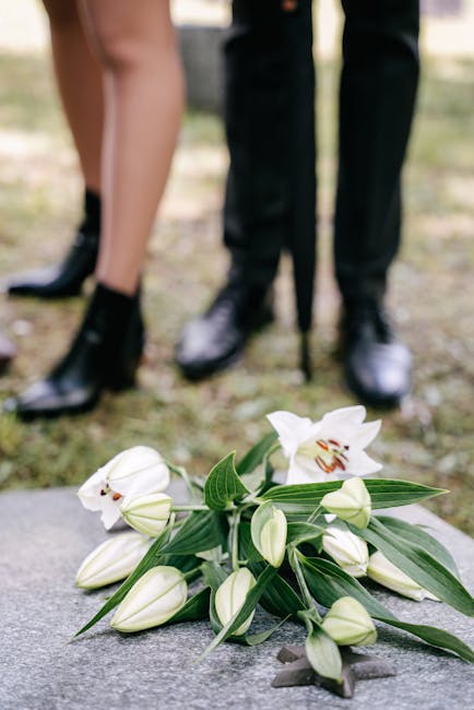 A bouquet of white lilies with large, open petals and prominent stamens, complemented by several closed lily buds and green elongated leaves, lies on a stone surface outdoors. The arrangement appears 