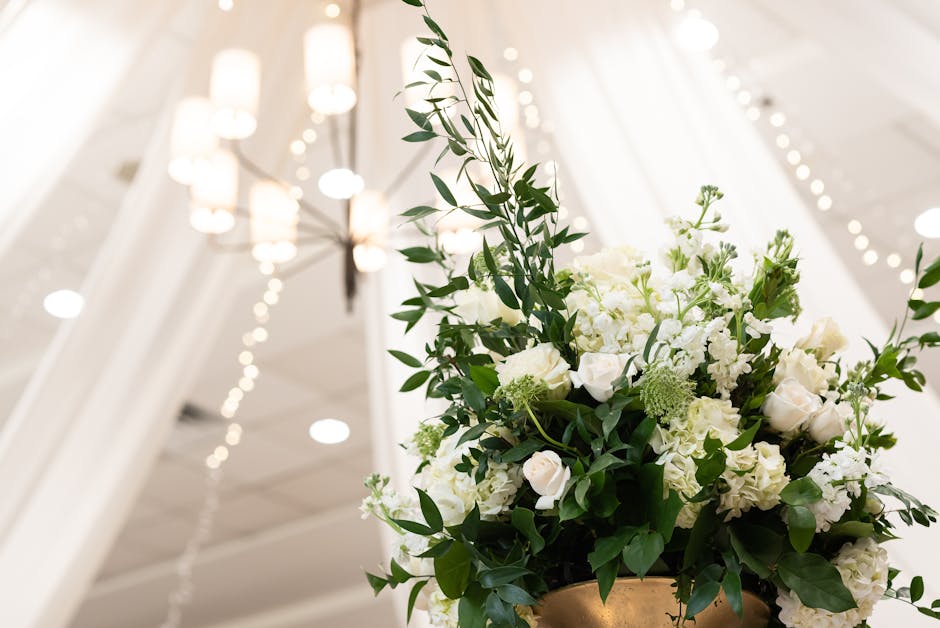 A floral arrangement featuring white roses, hydrangeas, and delicate greenery, arranged in a tall, elegant golden vase. The bouquet is placed on a table in an indoor setting with soft, ambient lightin
