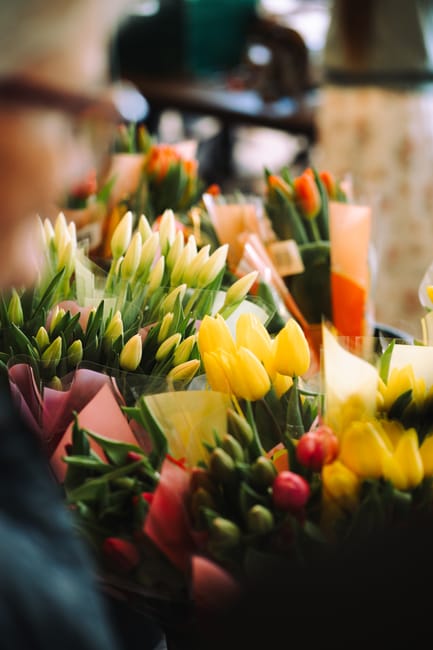 A vibrant display of fresh flowers, including white tulips, bright yellow tulips, and orange tulips, arranged in bouquets wrapped in pastel pink and yellow paper. The arrangements feature lush green s