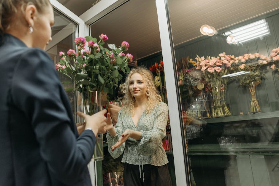 A woman with curly blonde hair, wearing a light-colored, pattern blouse, is inside a florist shop through a glass door, receiving a bouquet of pink roses with green leaves wrapped in brown paper from 