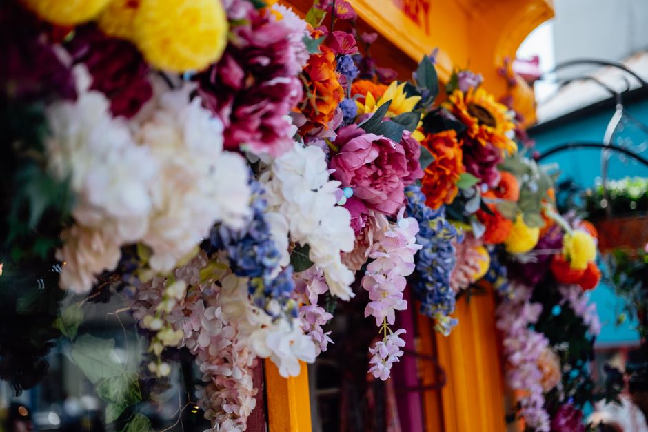 A vibrant floral display featuring a variety of fresh flowers draped along the side of a bright yellow vehicle. The arrangement includes pink peonies, white hydrangeas, orange marigolds, yellow sunflo