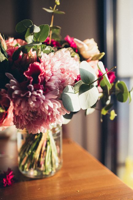 A vibrant floral arrangement placed in a clear glass vase on a wooden surface, featuring large pink peonies with ruffled petals, deep pink roses, and soft green eucalyptus leaves that add texture and 